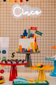 a table topped with cakes and donuts covered in legos next to a neon sign