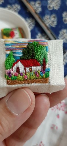 a hand holding a small piece of fabric with a house on it and trees in the background