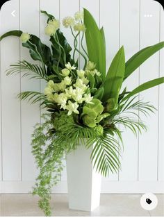 a white vase filled with lots of green flowers and greenery next to a wall