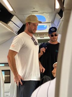 two men standing in the aisle of an airplane looking at something on his phone while wearing hats and sunglasses