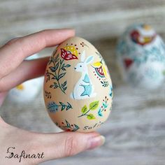 a hand holding an egg decorated with flowers and rabbits on it, next to another painted easter egg