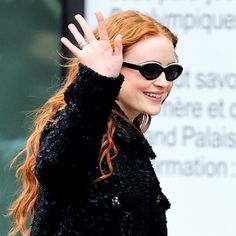 a woman with red hair and sunglasses waves to the side while wearing black clothing,