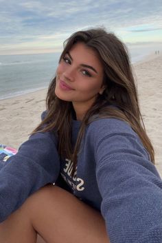 a beautiful young woman sitting on top of a sandy beach next to the ocean with her legs crossed