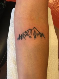 a small mountain tattoo on the left forearm and right arm, with pine trees at the top