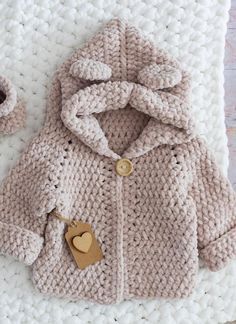 a crocheted baby coat and booties on a white blanket with a wooden heart