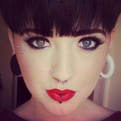 a woman with black hair and piercings on her nose is wearing red lipstick, white hoop earrings