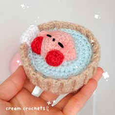 a hand holding a small crocheted pig in a basket with wings on it