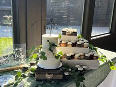 three tiered wedding cake with cupcakes and greenery on the table in front of large windows