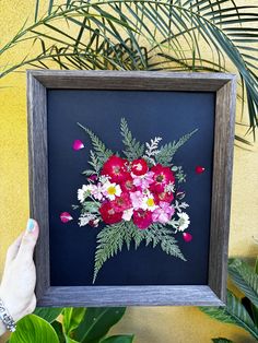 a person holding up a framed painting with flowers in the center and leaves around it