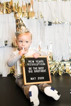 a little boy sitting on the floor holding a sign that says new years resolution be the best big brother