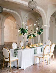 a table set up with white flowers and greenery in tall vases on top