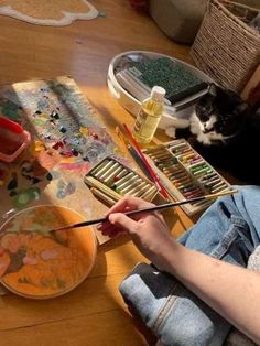 a person sitting at a table with paint and brushes in front of a cat laying on the floor