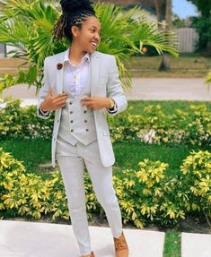 a woman wearing a white suit and tan shoes standing in front of some bushes smiling