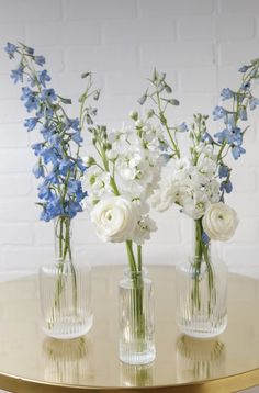 three clear vases with white and blue flowers in them on a round gold table