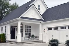 a white house with black shingles and two garage doors on the front porch, surrounded by greenery