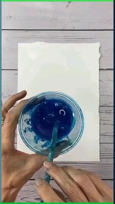 two hands are holding a glass with blue liquid in it and an empty piece of paper is on the table