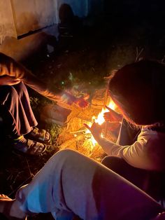 two people sitting around a campfire with their hands on the fire and looking at something