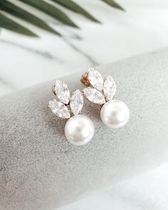 two pairs of pearl and diamond earrings sitting on top of a piece of fabric with palm leaves in the background