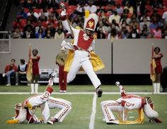 a man in red and white uniform jumping into the air on a football field with other people watching