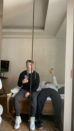 two men are sitting on a bed and one is taking a selfie in the mirror