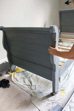 a woman is painting the headboard of a bed with gray paint and white sheets