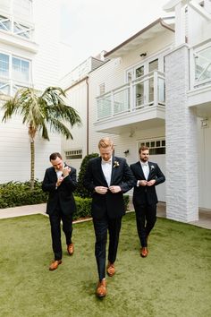 three men in tuxedos are walking through the grass near a building with balconies