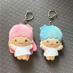 two key chains with small stuffed animals attached to them on a gray cloth surface, one is wearing a white dress and the other has blue hair