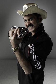 a man wearing a cowboy hat and holding a snake in his right hand while standing against a gray background