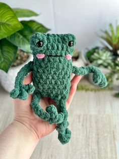 a small green crocheted frog sitting on top of a wooden table next to potted plants