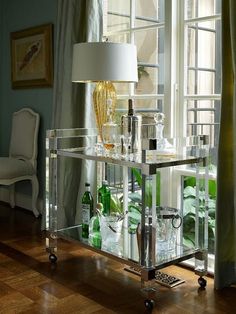 a glass bar cart with bottles and glasses on it sitting in front of a window