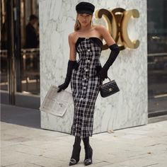 a woman in a black and white checkered dress is standing on the sidewalk with her handbag