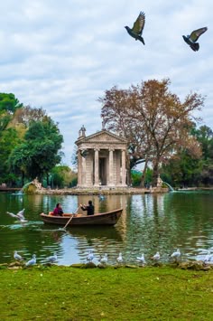 two people in a small boat on the water with birds flying over them and a gazebo in the background