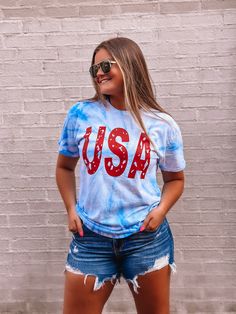 a woman in shorts and a tie dye usa t - shirt is standing against a brick wall