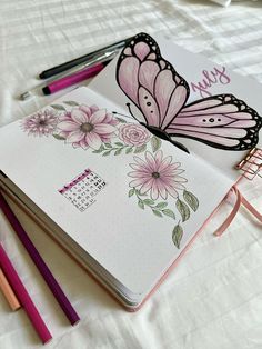 an open notebook with pink flowers and butterflies on it next to pencils, markers and a planner