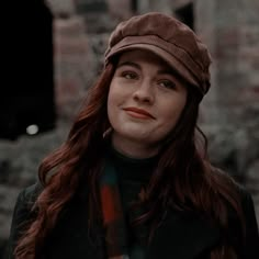 a woman with long red hair wearing a brown hat and black coat standing in front of an old brick building