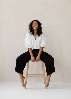 a woman sitting on top of a wooden stool in front of a white wall smiling