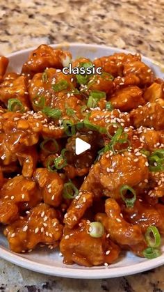 a white plate topped with chicken covered in sesame seeds and green onions on top of a granite counter