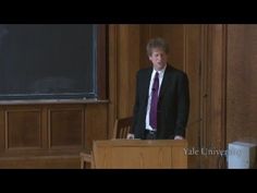 a man in a suit and tie standing at a podium with a chalkboard behind him