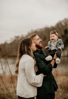 a man holding a baby while standing next to a woman in front of a body of water