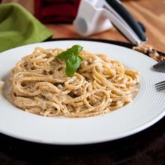 a white plate topped with pasta and garnished with green leafy basil next to a knife and fork