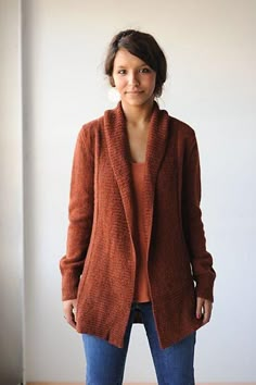 a woman standing in front of a wall wearing a brown cardigan sweater and jeans