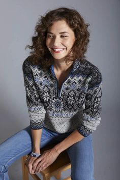 a smiling woman sitting on top of a wooden stool next to a white wall and wearing blue jeans