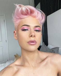 a woman with pink hair and makeup looks at the camera while sitting on a bed