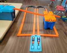 a toy train set on a wooden table with plastic containers and chairs in the background