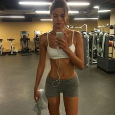 a woman standing in a gym looking at her cell phone and listening to headphones