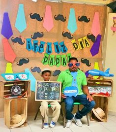 two men sitting in front of a bulletin board with fake mustaches on it and one man holding a sign