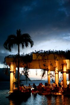 an outdoor dining area with palm trees on the water and people sitting at tables under umbrellas