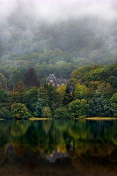 a house on the shore of a lake surrounded by trees and mist covered mountains in the background