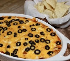 a white casserole dish filled with cheese and black olives next to a bowl of chips