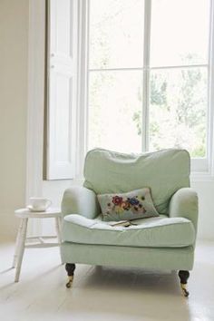 a light green chair sitting in front of a window next to a table with a vase on it